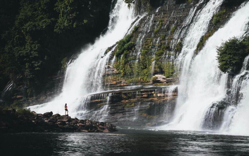 A woman stands on a rock outcrop at the base of Twin Falls, Rock Island, TN, a grand, double, cascade waterfall. 