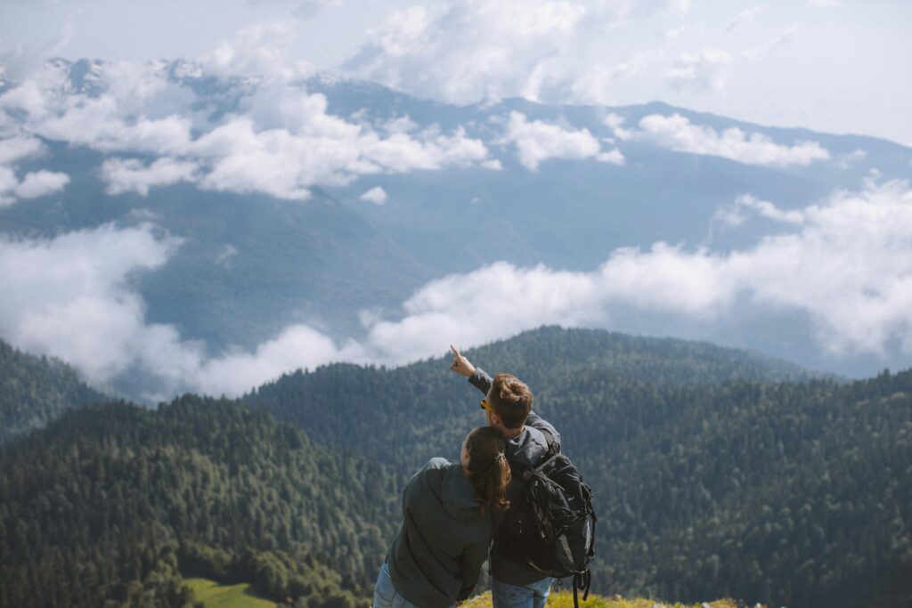 Couple stands looking at a vast mountain view. The young woman is leaning on the young man's shoulder, and the young man is pointing at something in the distance. Fluffy white clouds move between green, tree-covered mountains. 