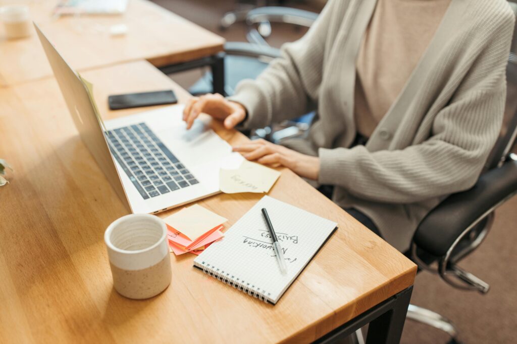 Shows woman in a beige shirt and gray cozy cardigan at a butcher block desk working on a laptop with a phone, coffee mug, and notepad nearby. 